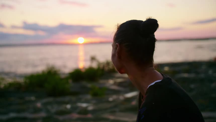 Woman looks out to sea watching sun hover on horizon in red tinted sky
