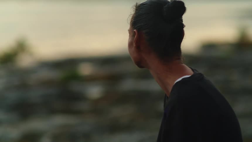 A woman sits on a large stone while gazing out at the ocean under an evening sky