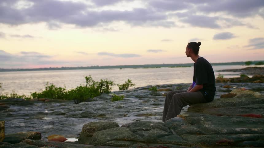 A man sits on a rock on a shoreline looking at the body of water