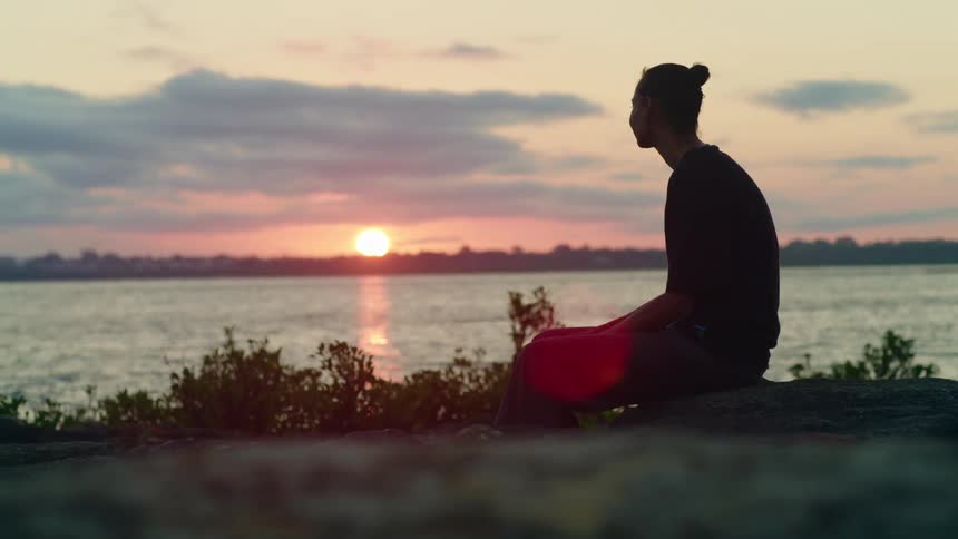 A woman sits on a stone on a lake shore as she watches the setting sun on the horizon