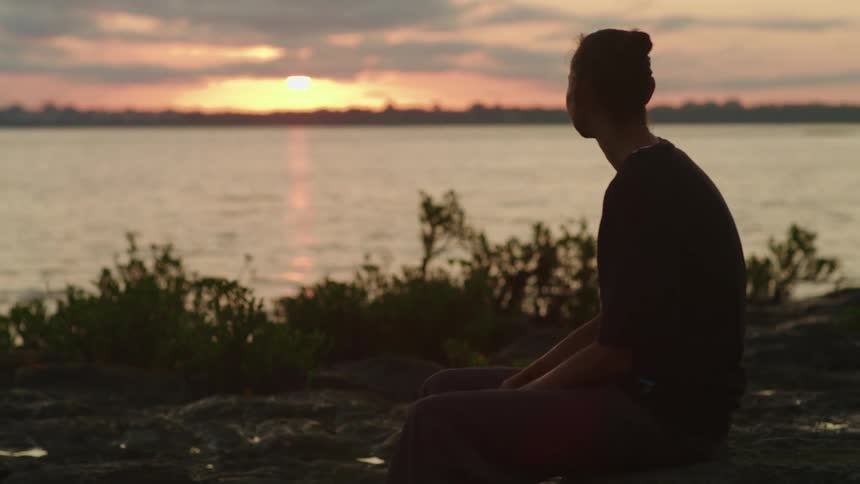 An aboriginal woman sitting near a lake shore watching the sun set