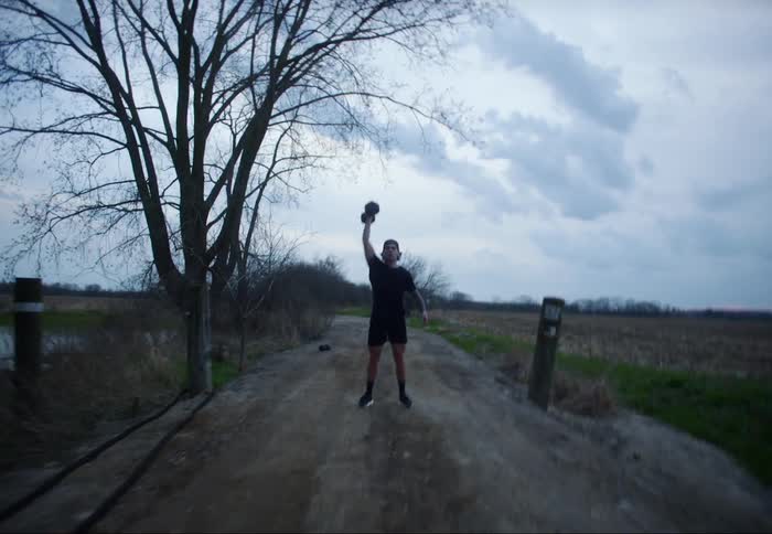 Man with a cap a black t shirt and shorts works out with a dumbbell on a dirt road in the countryside