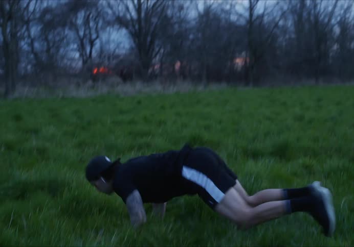 Man with a cap and a black t-shirt does burpees on the green grass in a field at dusk