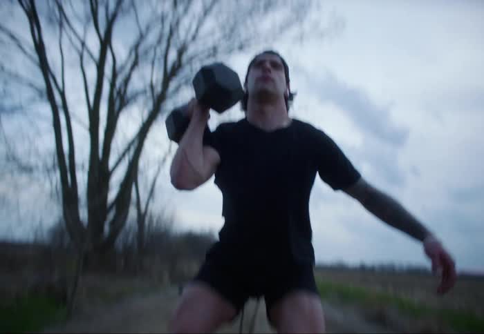 A man lifts a heavy dumbbell while doing squats on a dirt road in the countryside on a cloudy evening