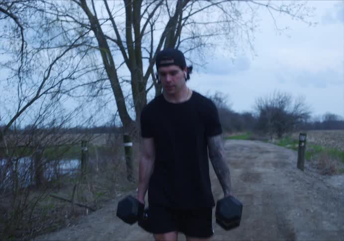 Man with a cap earphones and a black t-shirt carries two dumbbells on a dirt road on a cloudy day