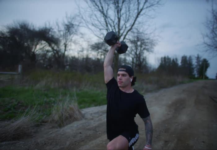 A man holds a dumbbell up over his head with one arm while doing lunges down a country dirt road