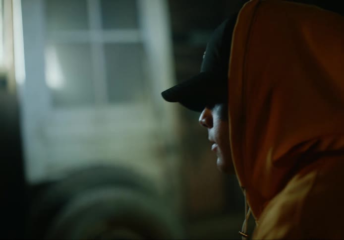 Man wearing hoodie and baseball cap leans forward in a dim room