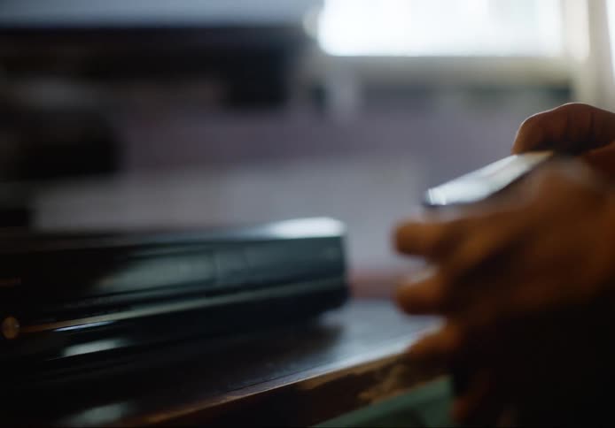 Person wearing long sleeved shirt picks vhs tape from on top of vhs player and reads label