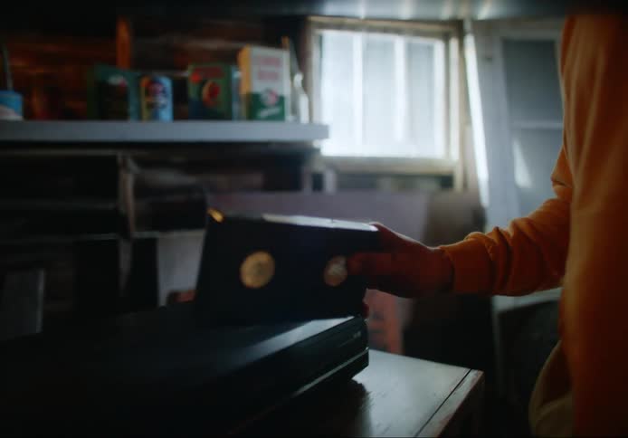 A man raises from his seat to approach a television set and pick up a video cassette on top