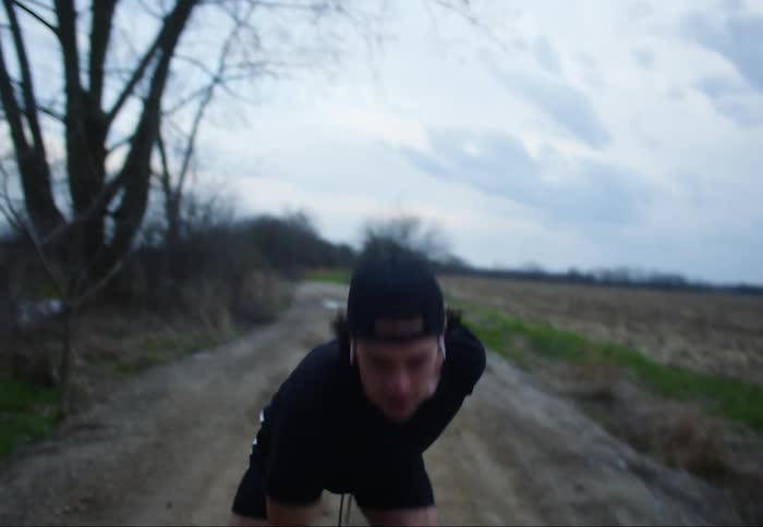 A man is vigorously lifting a dumbbell while standing on a dirt road next to farmland on a cloudy fall day