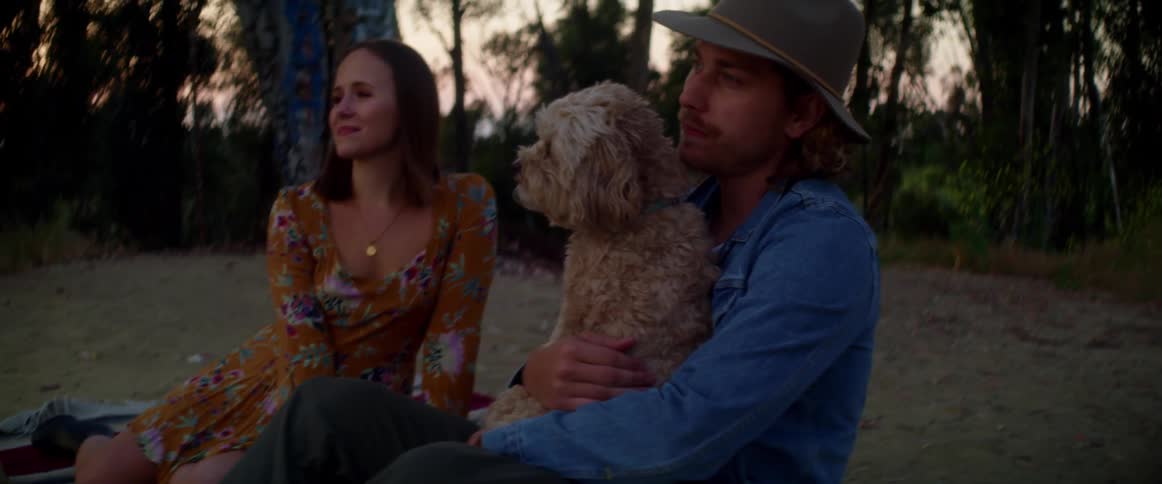 A man holds and pets a dog in his lap while sitting on the ground beside a woman at sunset