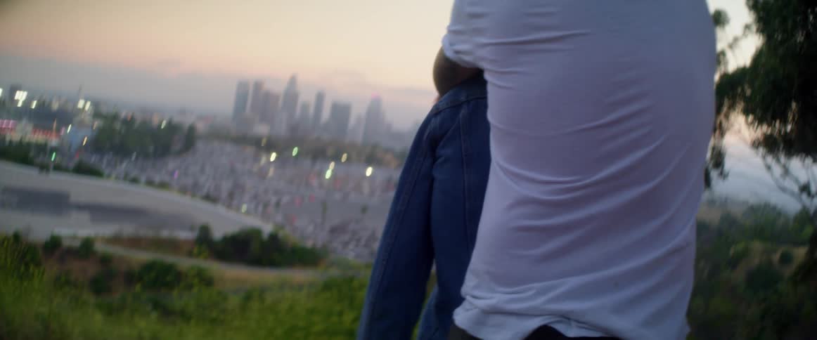 A man puts his jacket over a womans shoulder and then stands behind her and hugs her as they look at the city skyline