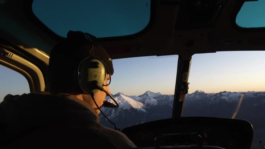 A man pilots a helicopter above a snow capped mountain range during the day
