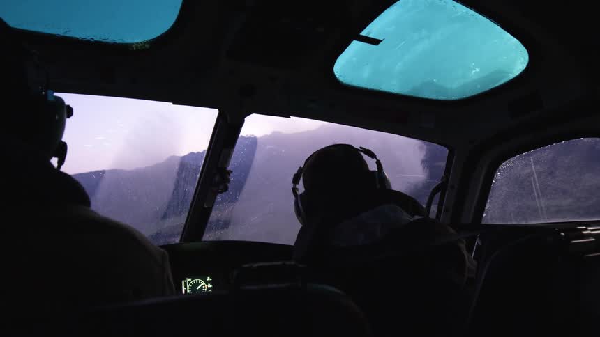 A helicopter pilot and his passenger chat as they fly over a mountain range