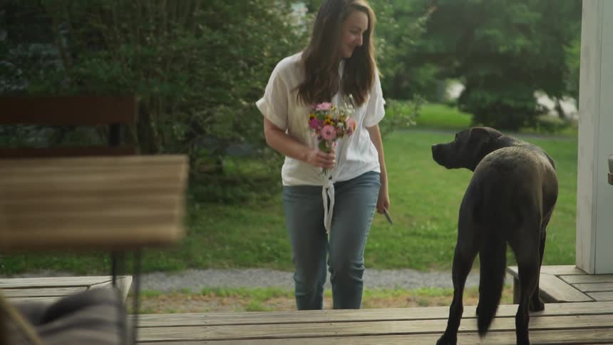A young woman carrying a bouquet of flowers walks past her dog and up her porch steps