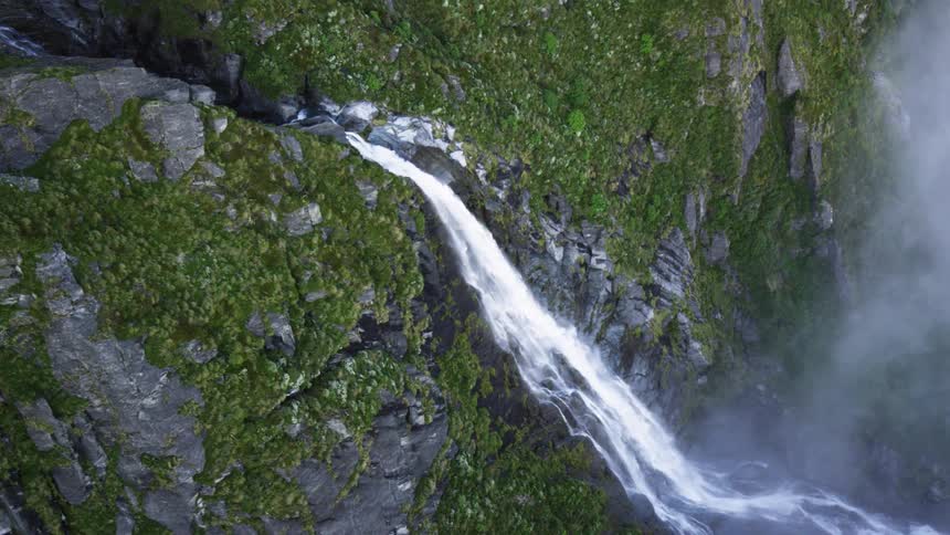 Water falls from a rocky cliff covered in thin vegetation into a misty valley