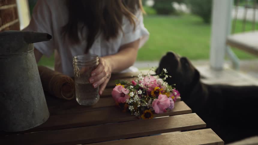 A woman sits on a porch pouring a glass of water and her dog comes over for attention