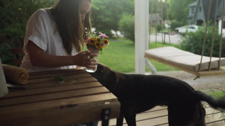 A woman sitting on her front porch lets her dog smell the freshly cut flowers in a vase