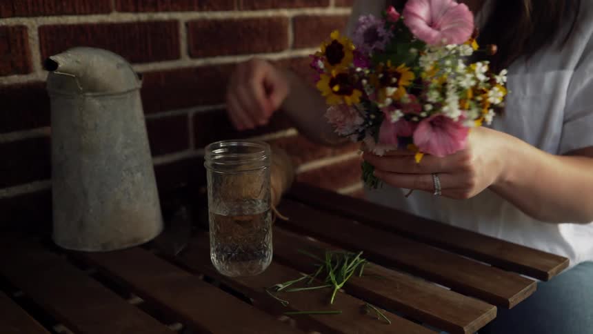 A woman uses a pair of scissors to trim the stems of a bouquet of flowers before placing the bouquet in a jar half filled with water