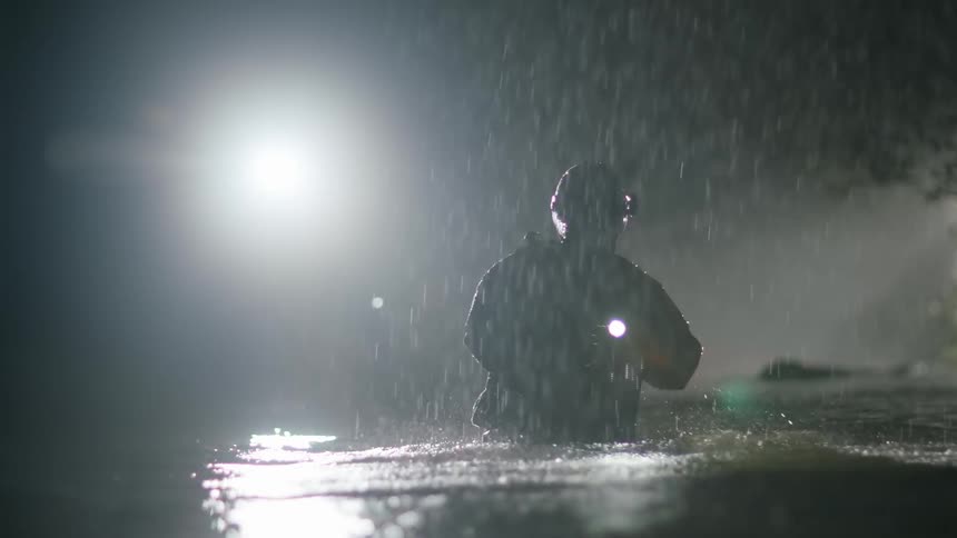 A first responder wades in to a lake in the pouring rain and searches with his flashlight