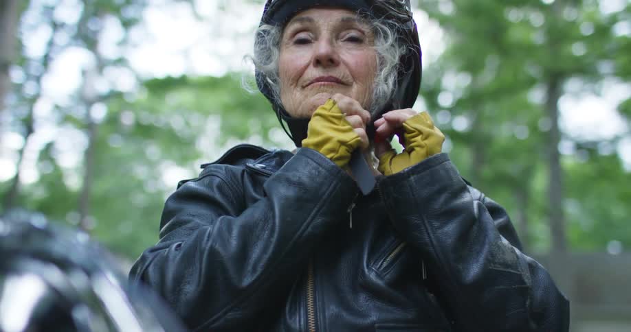 Elderly woman with a leather jacket and gray hair puts on a helmet before riding the motorcycle