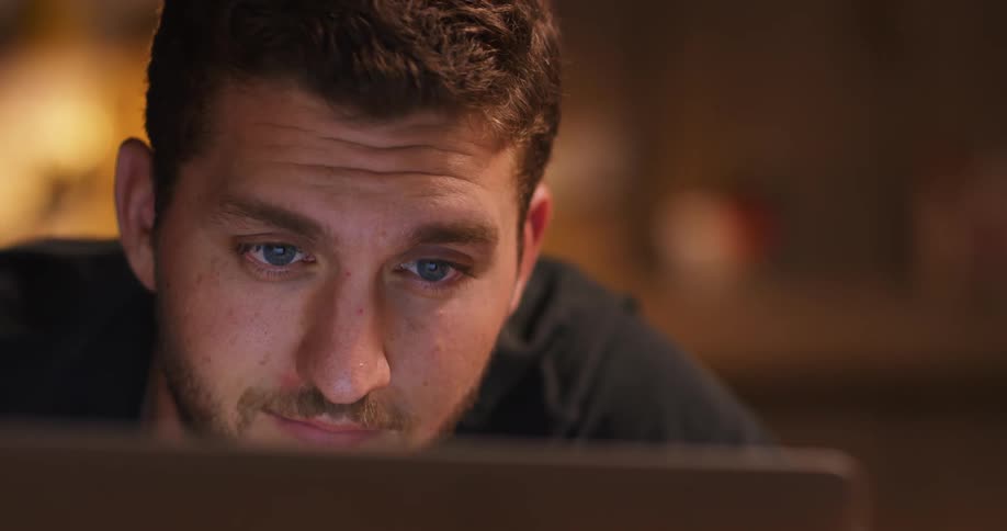 Man with a beard and a black t shirt smiles slightly and uses a laptop in a room