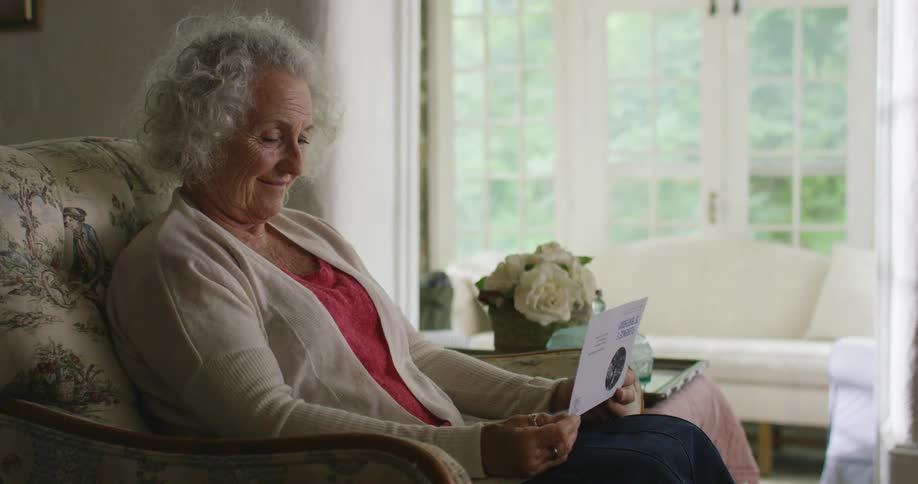 Elderly woman with grey hair sits in an armchair and reads a letter smilingly in the living room
