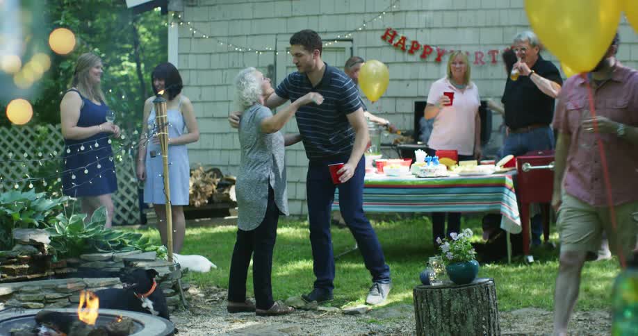 A young man hugs his grandmother at a backyard birthday party with a fire pit and yellow balloons
