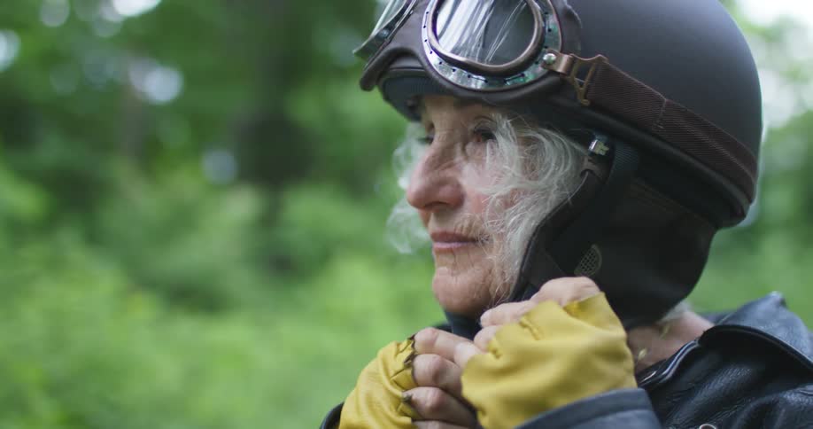 Elderly woman with a black leather and grey hair jacket puts on a motorcycle helmet