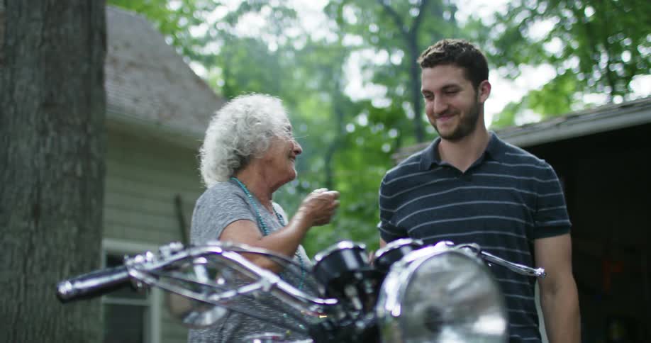 A man pulls a cover off a motorcycle and his mother covers her mouth and smiles and hugs him