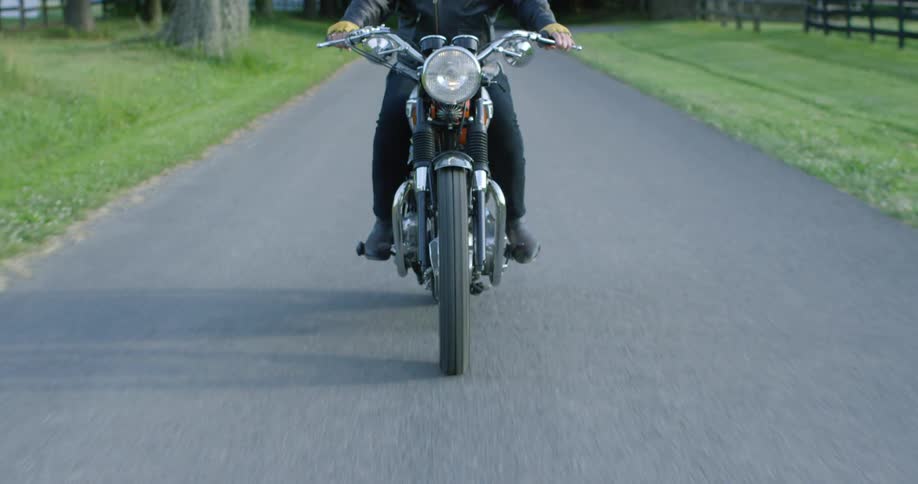 A biker in a leather jacket rides a motorcycle down a narrow road during the day