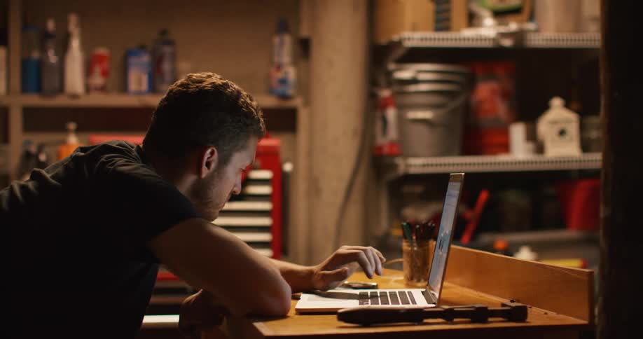 Man with a black t shirt sits at a table and uses a laptop in a room