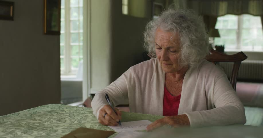 Elderly woman with grey hair sits at a table writes a letter in the living room at home