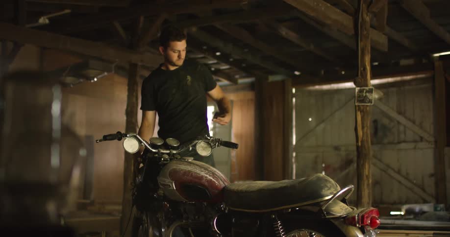 A man standing in front of a dusty motorcycle in a wooden building removes his smartphone from his back pocket to look at the screen