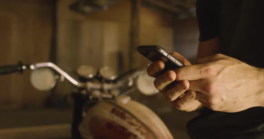 Man with a black t shirt leans against a motorcycle and uses a smartphone