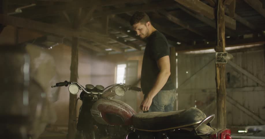 A man lifts a tarp to reveal a dusty motorcycle which he tries to clean by blowing the dust off