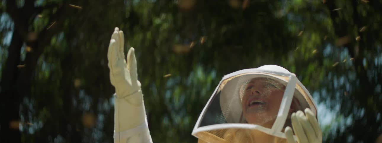 A bee keeper in personal protective equipment watches excitedly as bees fly around her