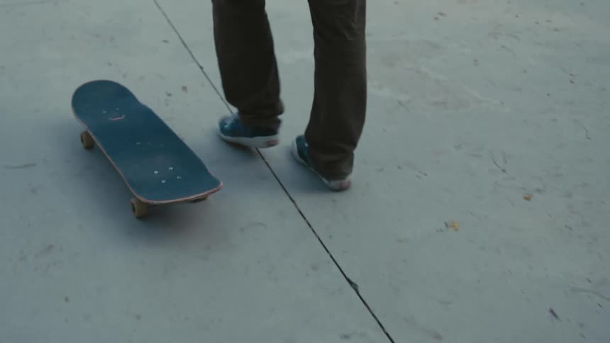 A skateboarder walks along a walkway as their skateboard rolls beside them
