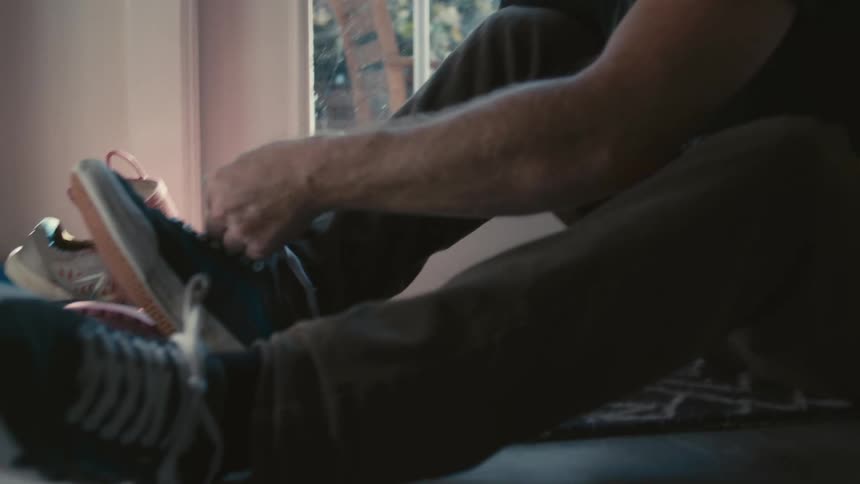 A middle aged man sits on the floor while he puts on and ties his shoes