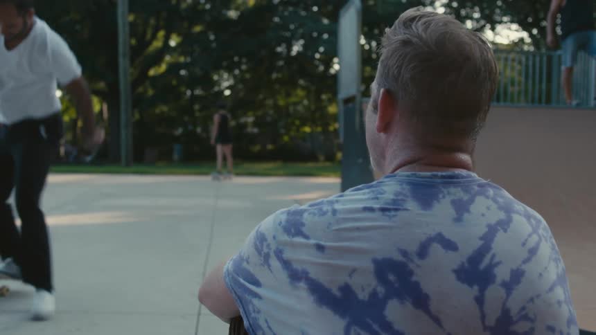 A man sits on the ground watching others skateboard in a public park