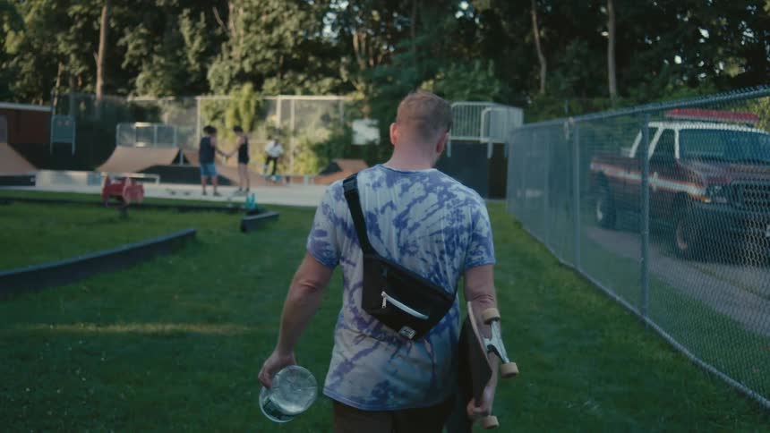 Man carrying skateboard walks across lawn toward skate park