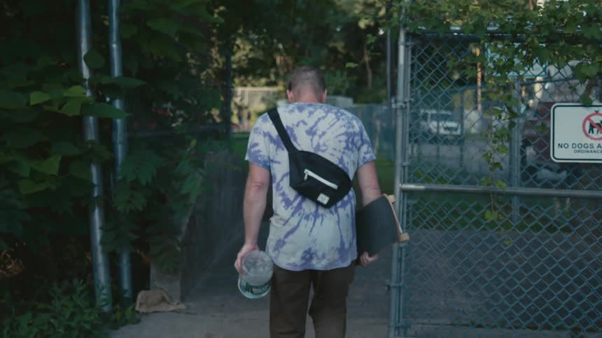 A man carries a skateboard and large water jug into an outside fenced area