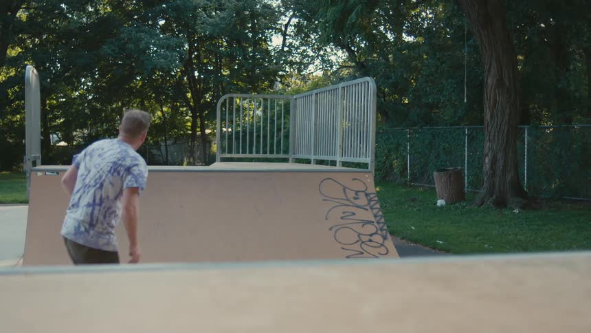 A man in a skateboard park skates up and down ramps and then jumps off his board and runs up a ramp and lies down