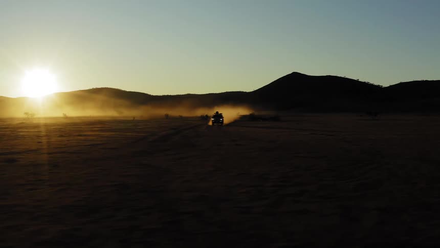 A vehicle races across a dusty field at sunset