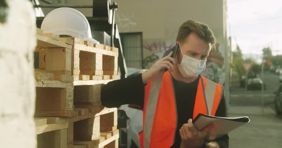 A construction worker holds a notebook and talks on the phone outside standing next to a stack of pallets