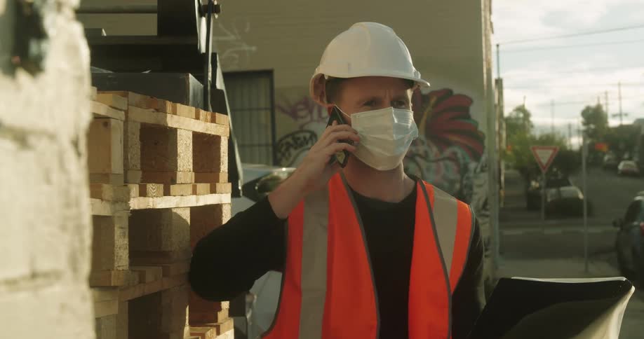 Man wearing hard hat and face mask consults notebook while talking on mobile phone near building as traffic passes on nearby street