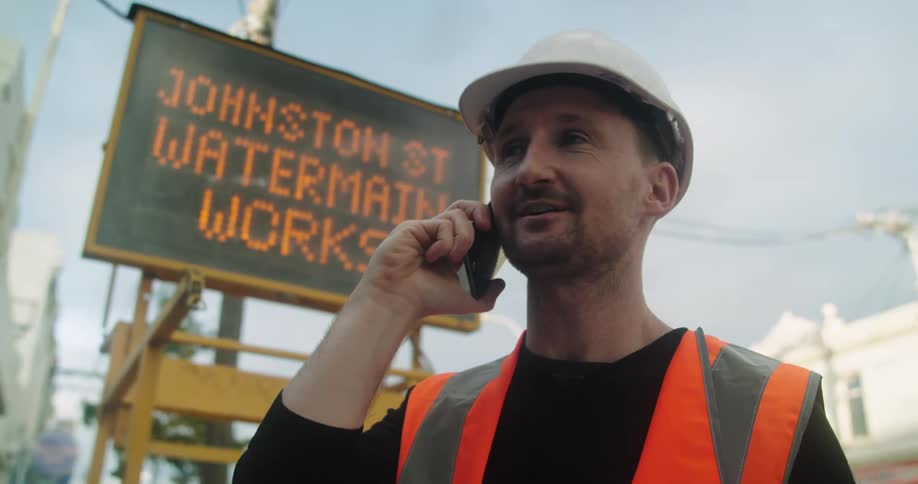 A man with a hard hat and a vest talking on a smart phone with a sign in the background