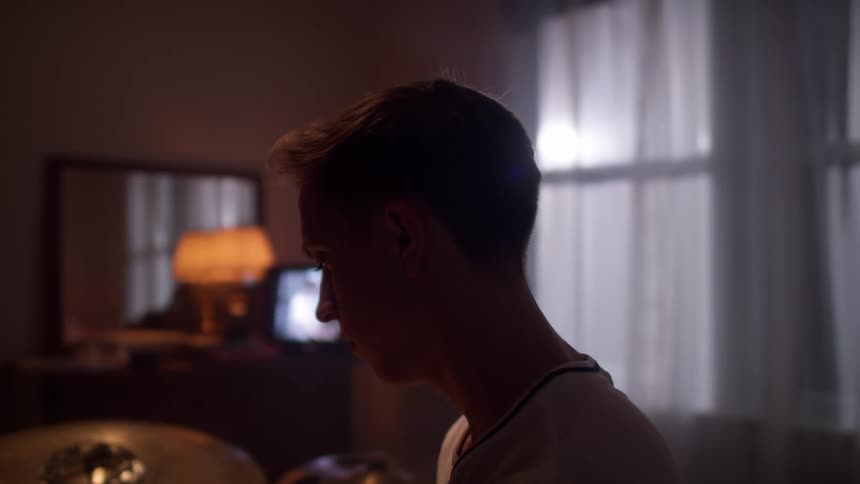 A man sits at a drum set in his bedroom with a television playing in the background