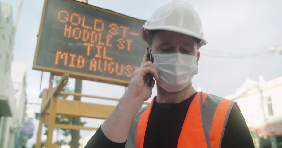 A construction worker looks at plans and talks on his cellphone while standing on a city sidewalk in front of a traffic sign