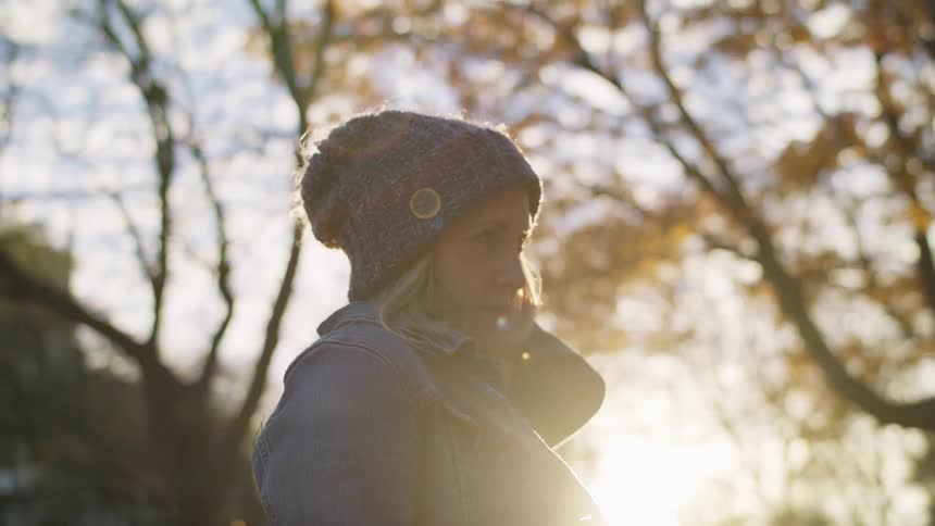 A woman wearing a jean jacket and knit hat stands under autumnal trees
